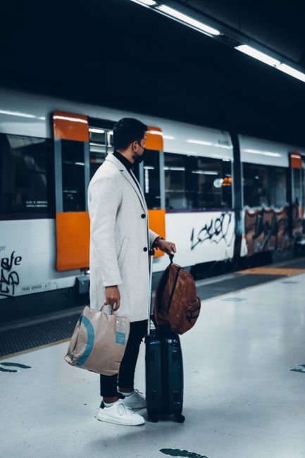 A woman with long dark hair, dressed in a black coat, stands on a platform at Catford Bridge Station, holding a black tote bag with social media hashtags, while facing away from the camera. The statio