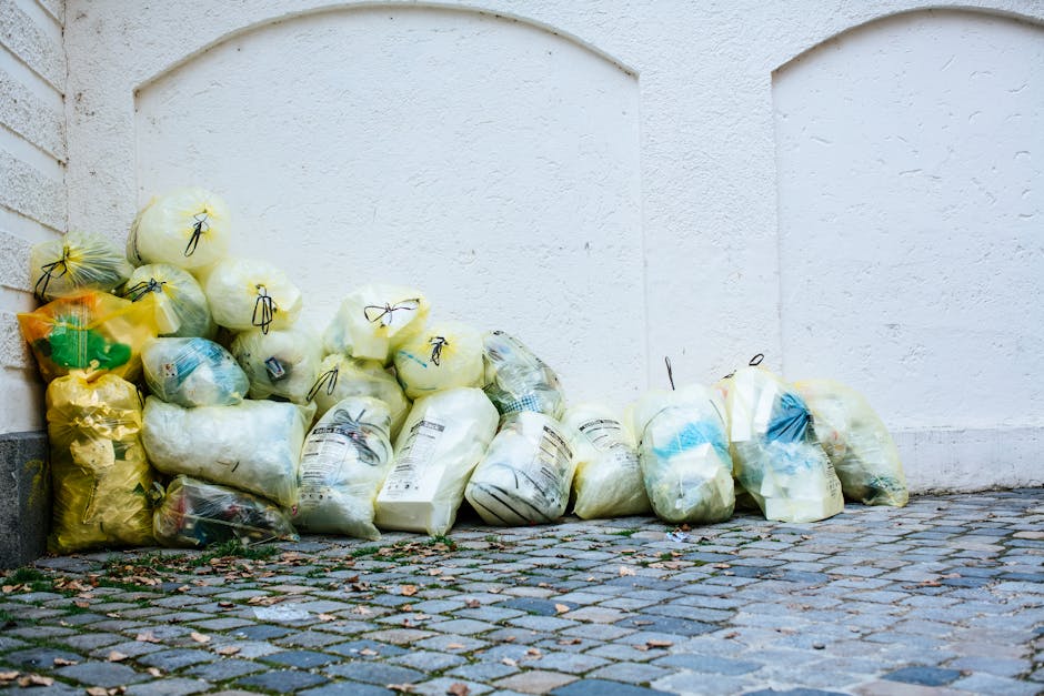 A pile of tightly tied black, yellow, and transparent garbage bags filled with waste, placed on a cobblestone pavement against a white exterior wall with a curved arch detail. The area appears clean b