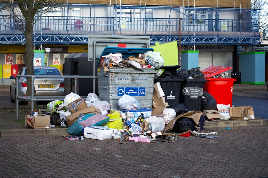 A pile of tightly tied black, yellow, and transparent garbage bags filled with waste, placed on a cobblestone pavement against a white exterior wall with a curved arch detail. The area appears clean b